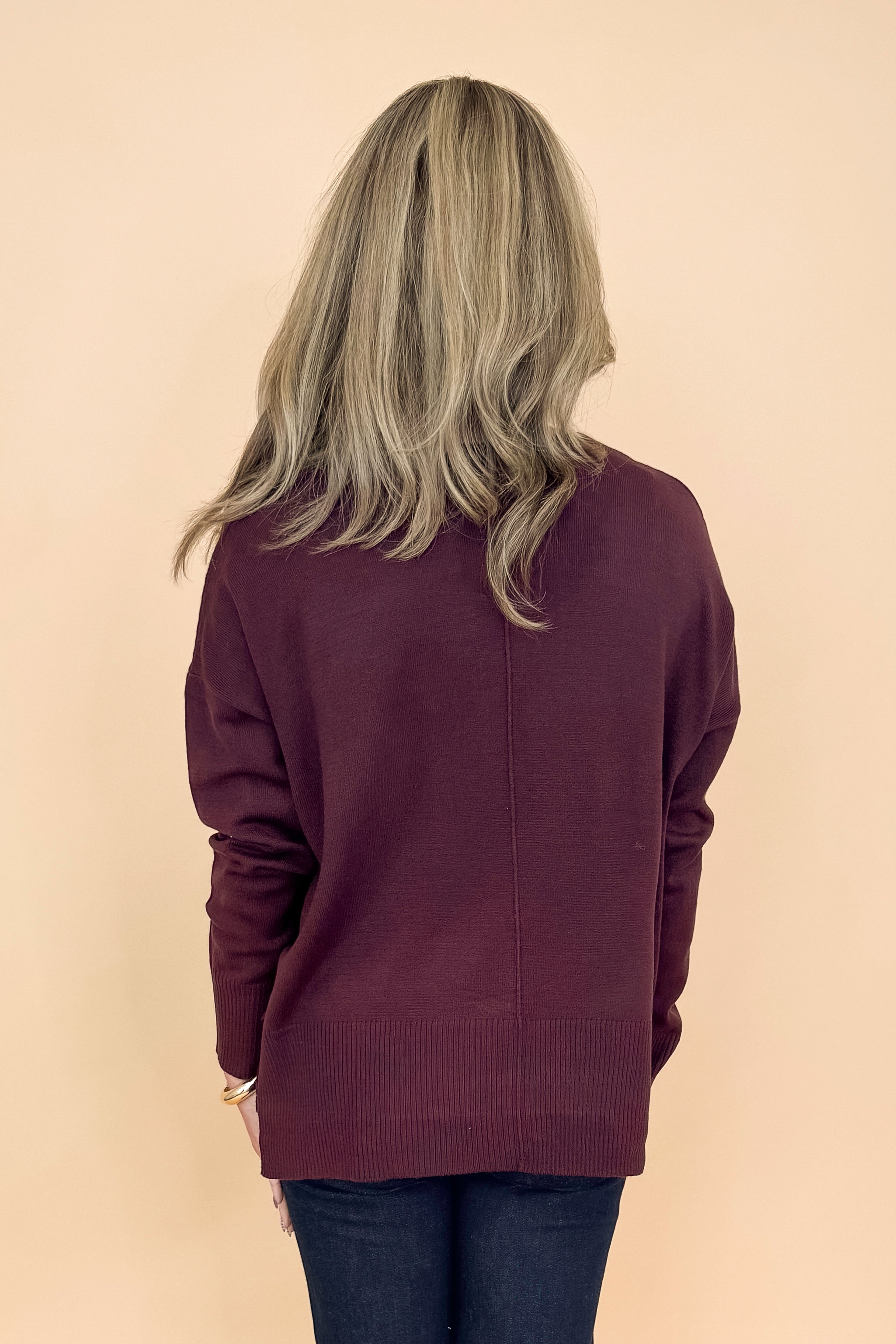 Back view of woman in a studio wearing Wine Center Seam Round Neck Sweater and jeans with a tan background. 