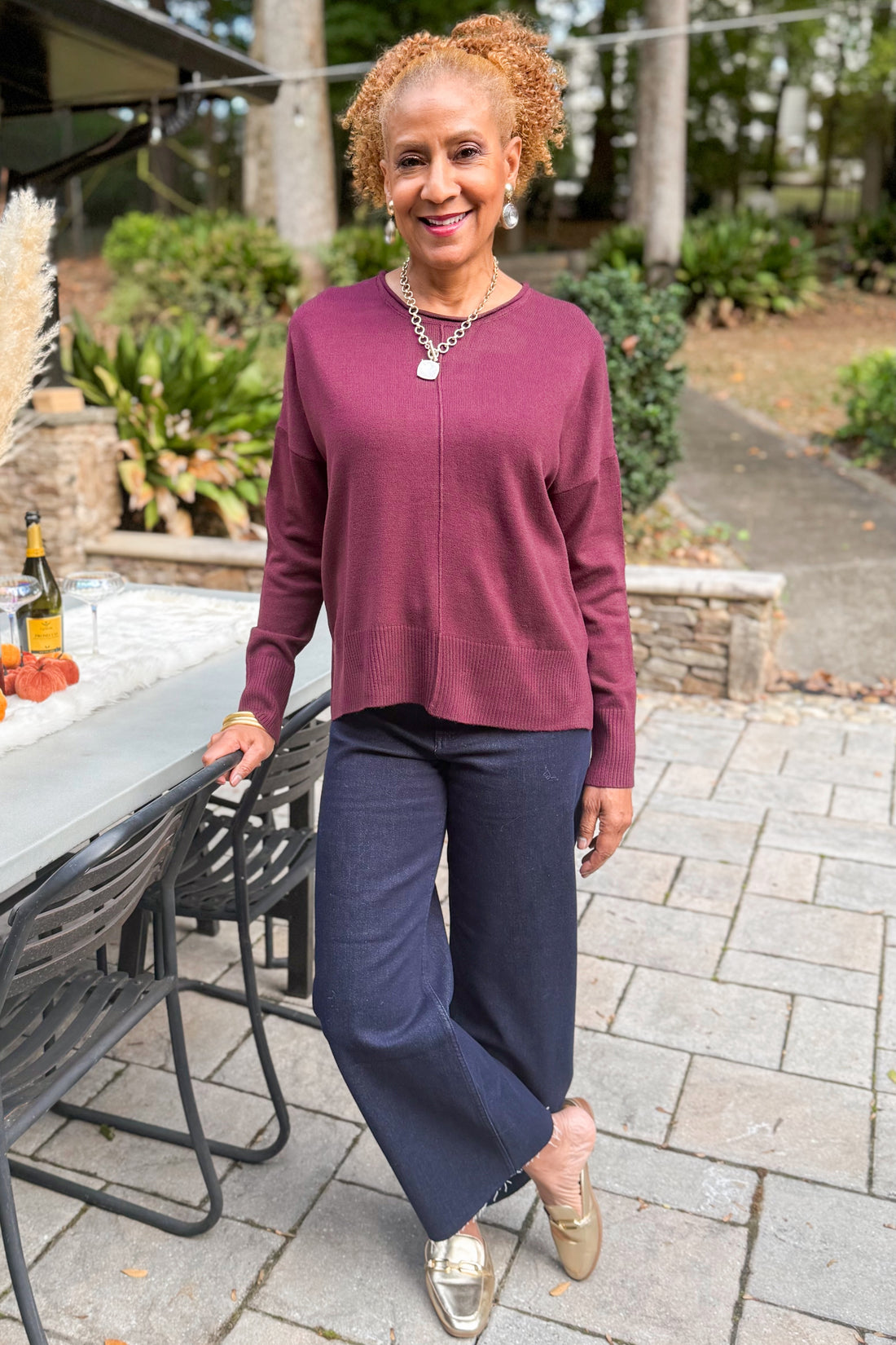 Woman outside wearing Wine Center Seam Round Neck Sweater and jeans by a table.