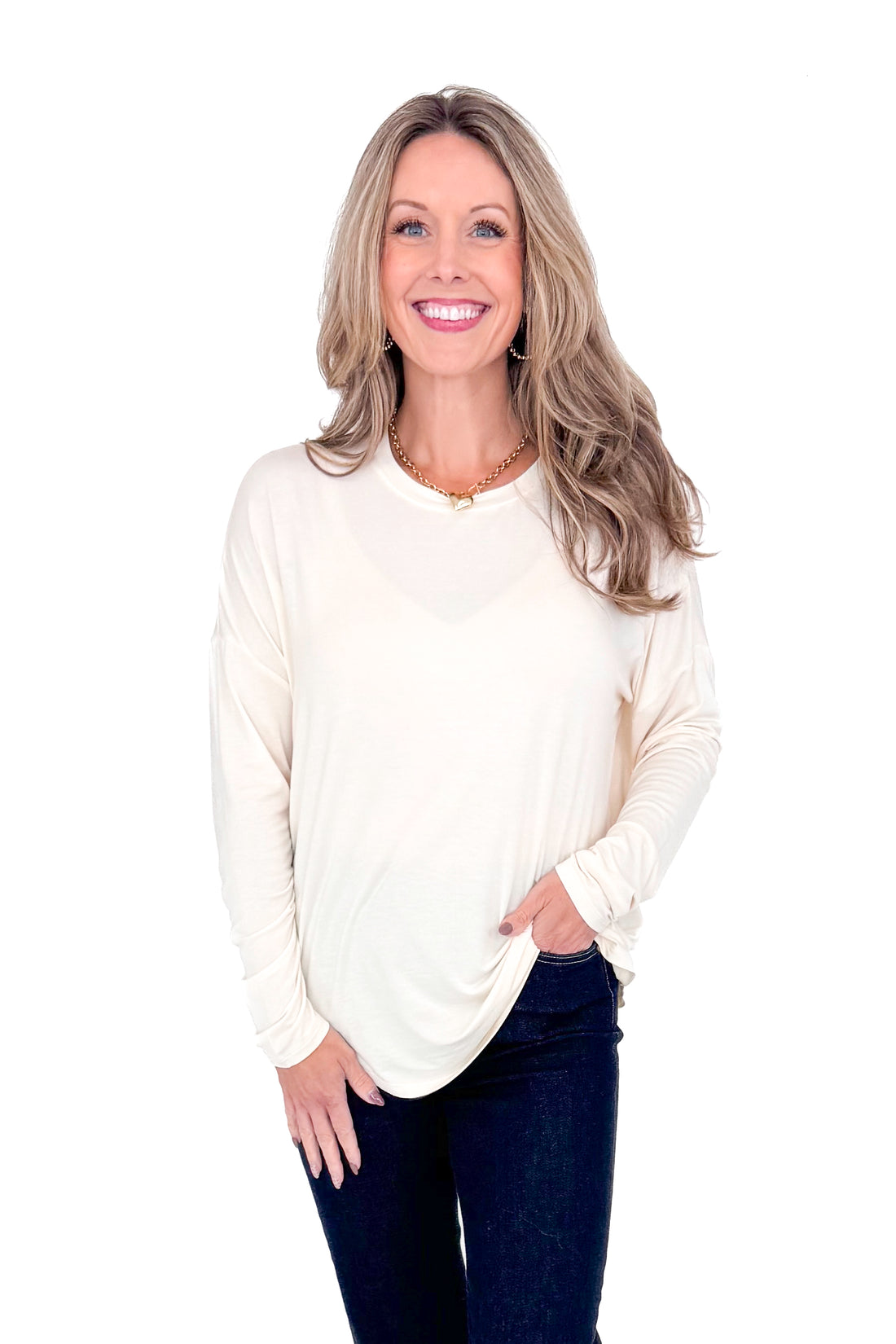 Front view of woman with cream long sleeve curve hem top and blue jeans against white studio background.