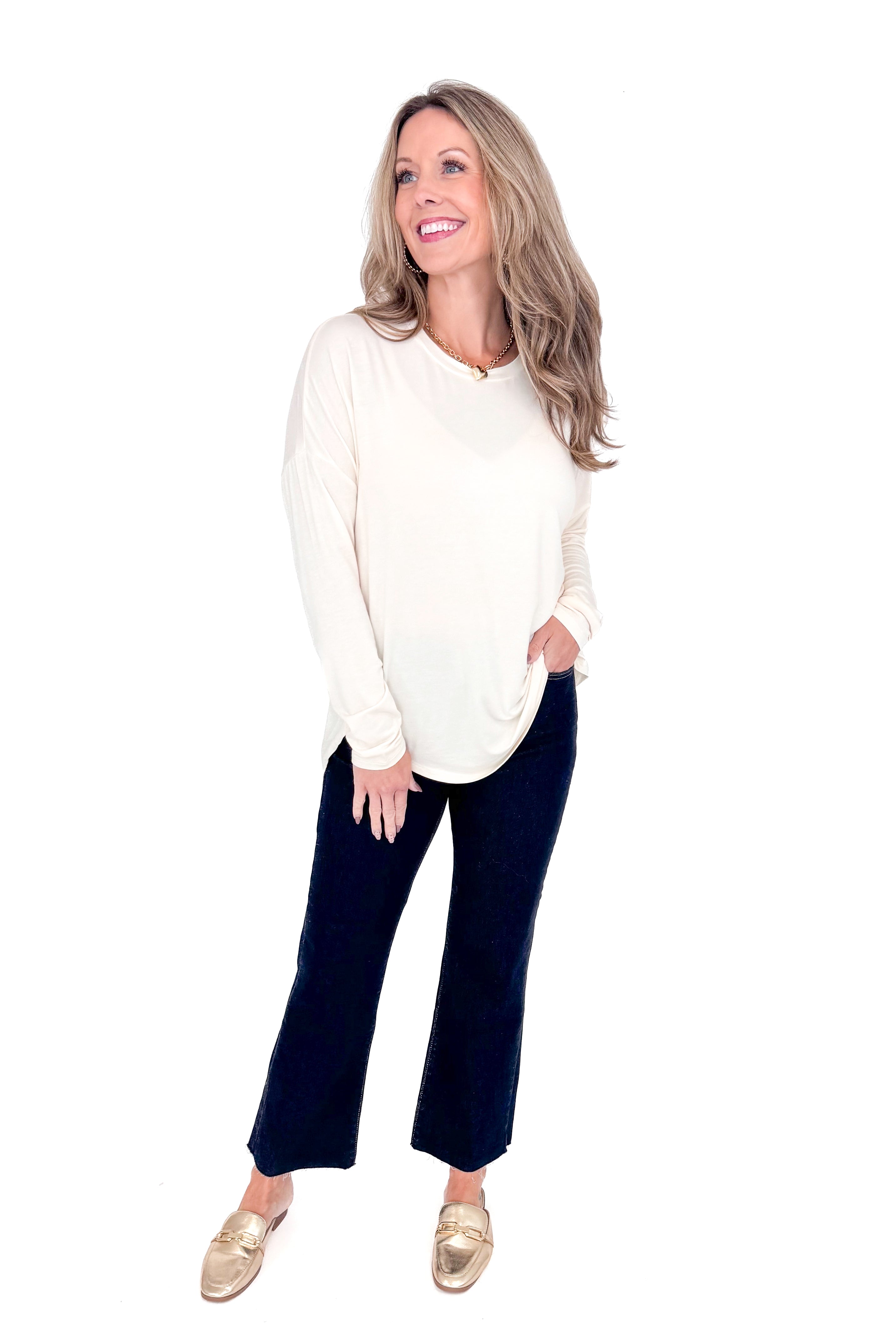 Front view of woman with cream long sleeve curve hem top and blue jeans against white studio background.