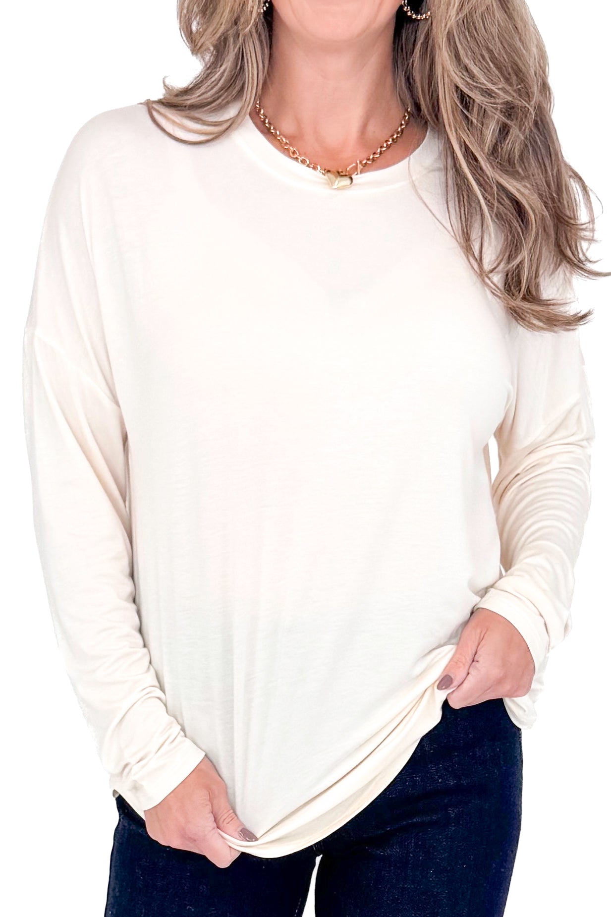 Front view of woman with cream long sleeve curve hem top and blue jeans against white studio background.