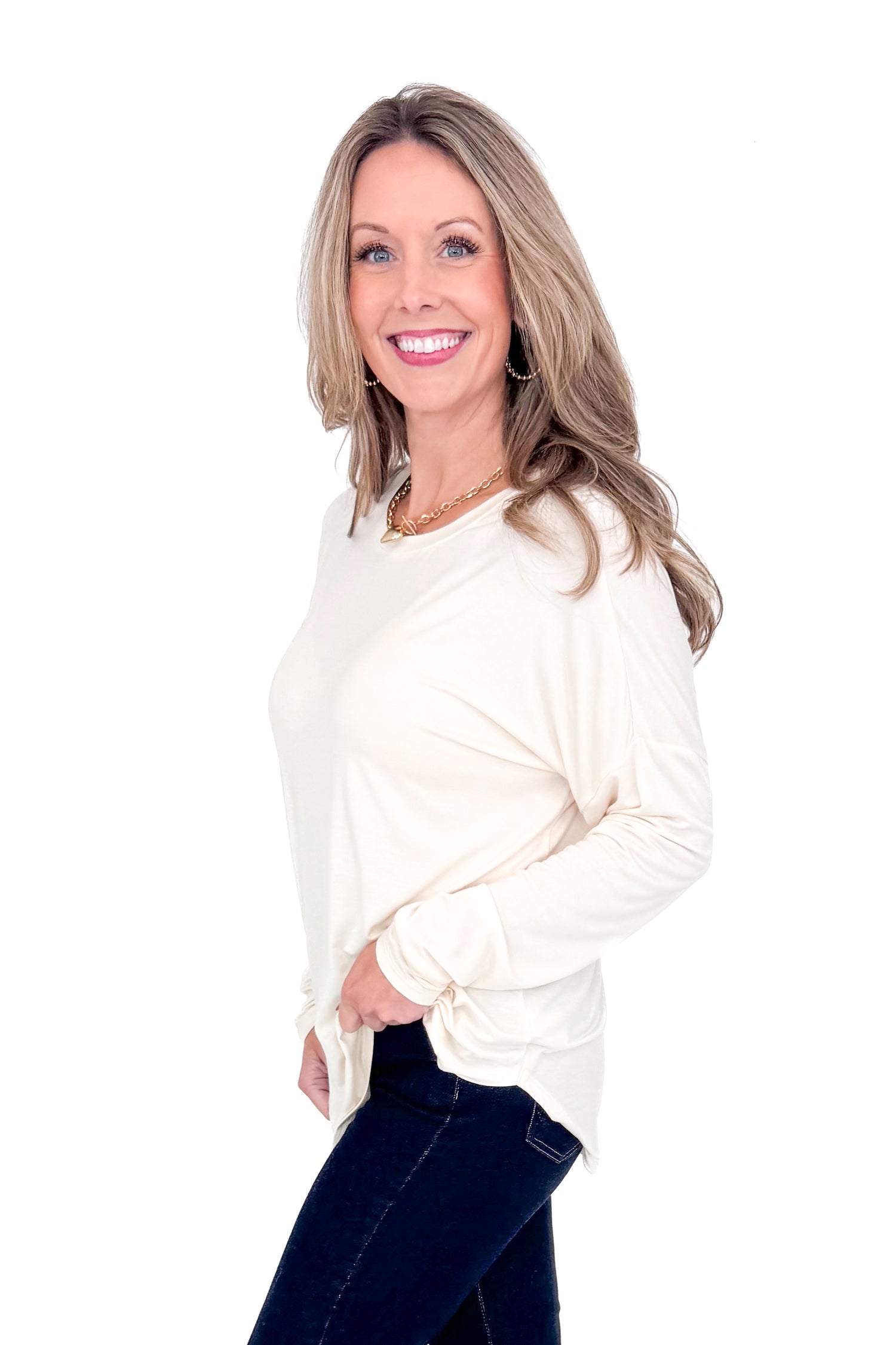 Side view of woman with cream long sleeve curve hem top and blue jeans against white studio background.