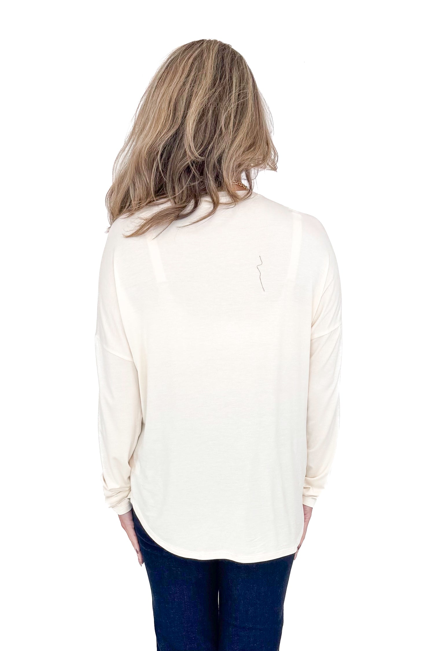 Back view of woman with cream long sleeve curve hem top and blue jeans against white studio background.