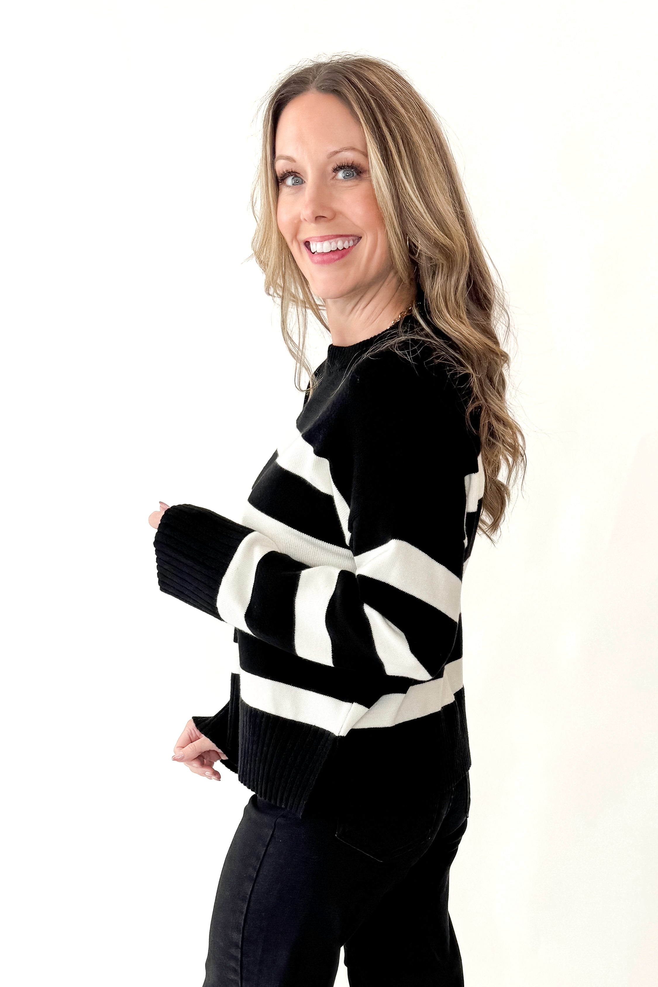 Side view of woman wearing Black and Cream Striped Crewneck Sweater with jeans against white studio background.