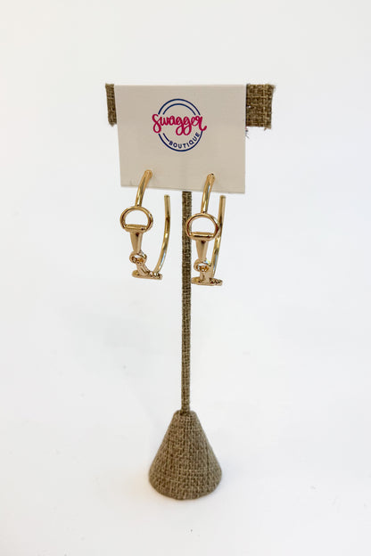 A pair of gold horse-bit style hoop earrings displayed on a textured stand with a branded card that reads &