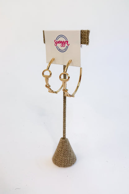 Gold-tone horsebit hoop earrings displayed on a burlap-covered stand with a branded card that reads &