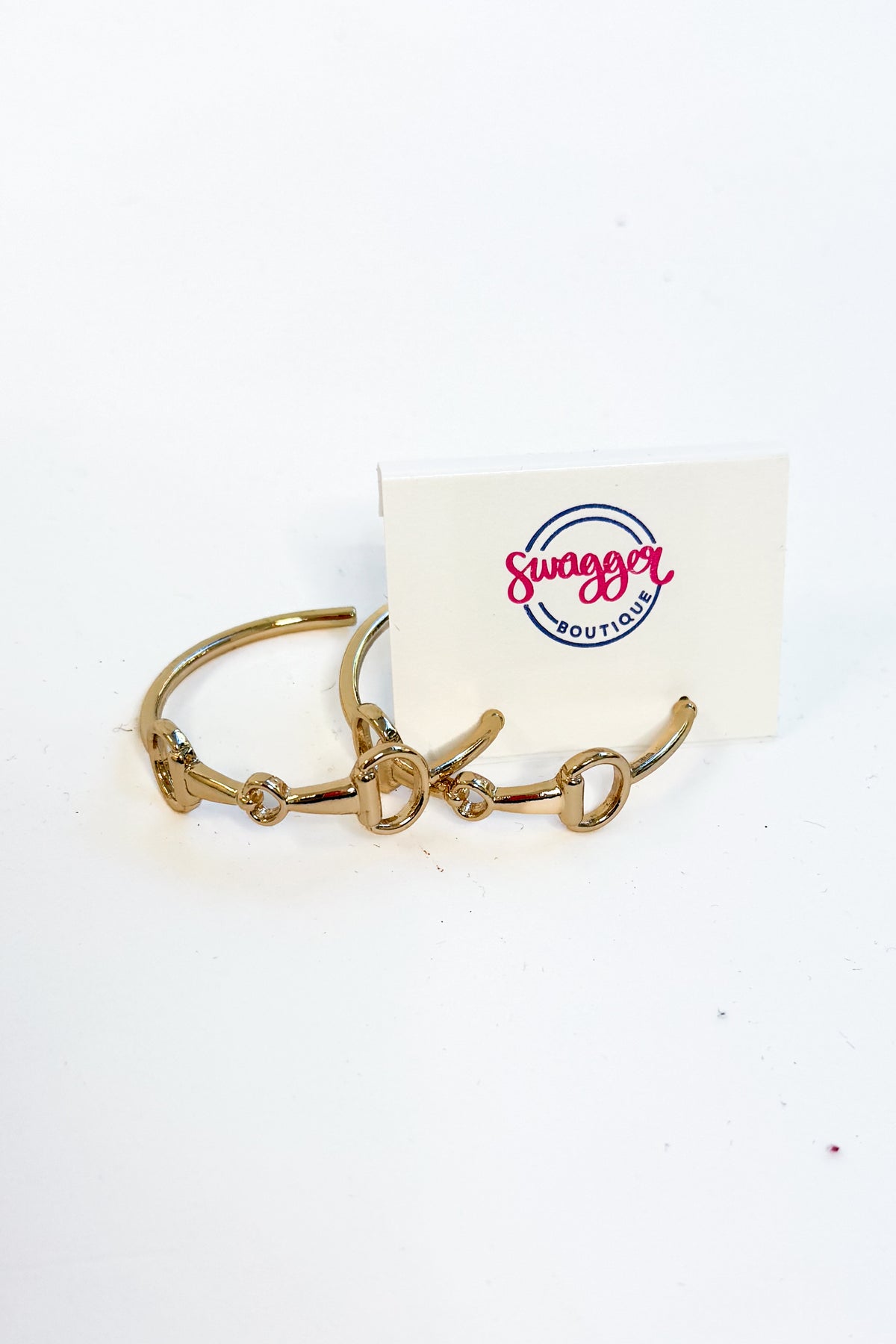 A pair of gold-toned metal cuff earrings with a horse-bit motif, displayed next to a white card featuring the &