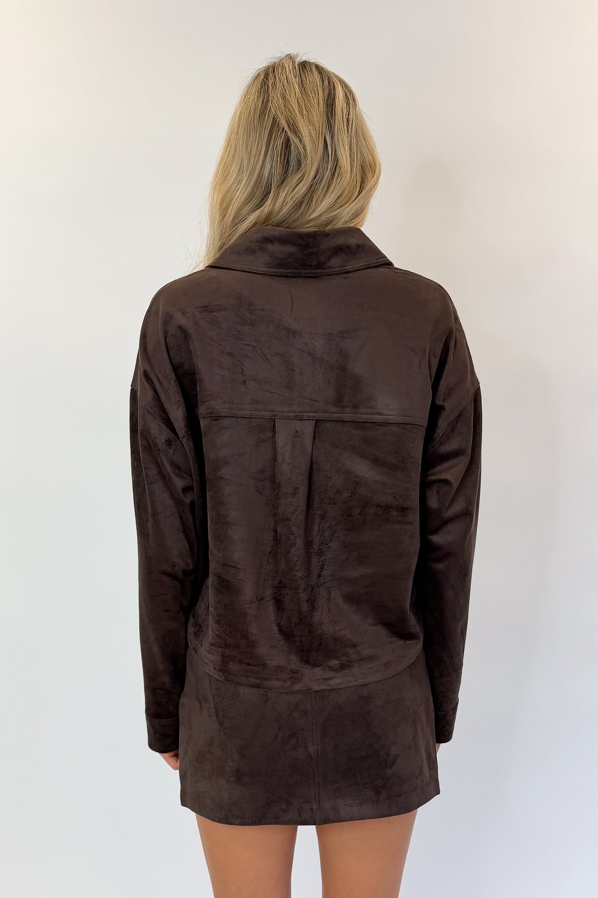 Back view of woman wearing Swagger Boutique’s All Day Cropped Suede Jacket and Perri Suede Mini Skirt by Z Supply In Dark Chocolate against white studio background.