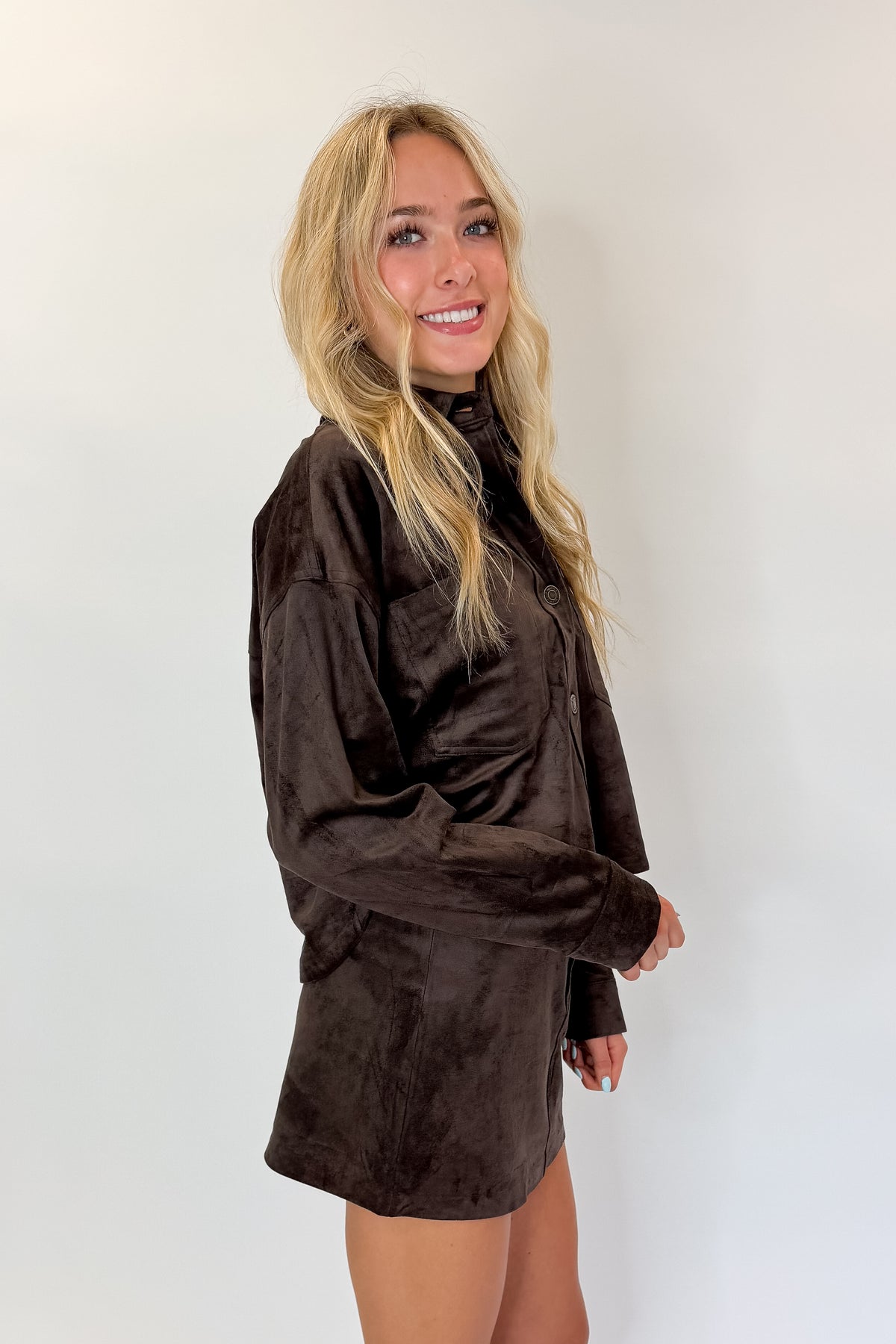 Side view of woman wearing Swagger Boutique’s All Day Cropped Suede Jacket and Perri Suede Mini Skirt by Z Supply In Dark Chocolate against white studio background.