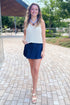 Front view of woman wearing Swagger Boutique’s Dark Denim Wrap Skort with White V-Neck Knitted Tank walking outside.