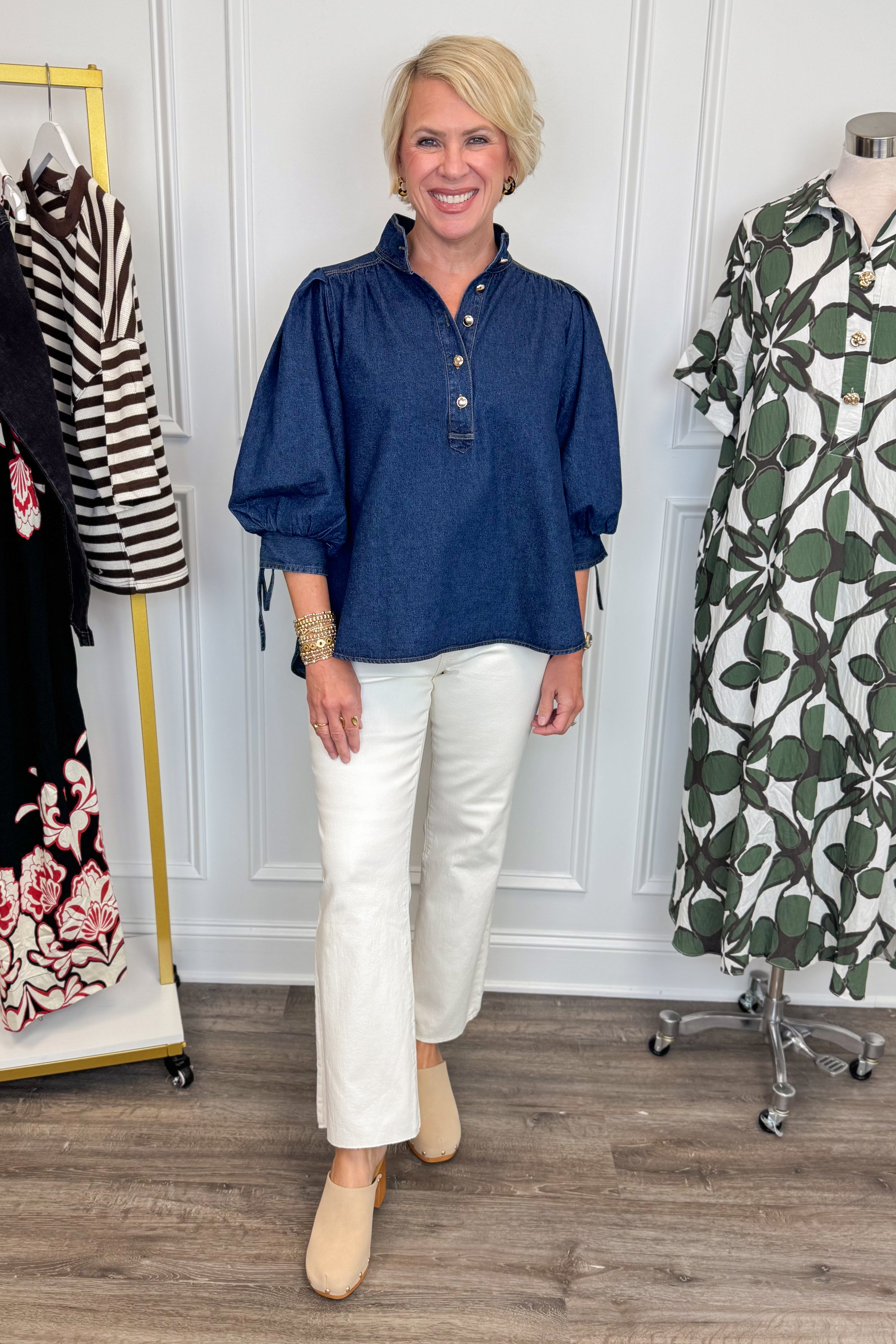 Front view of woman wearing Swagger Boutique’s Dark Wash Puff Sleeve Denim Top by Entro with jeans with clothing hanging in the background.
