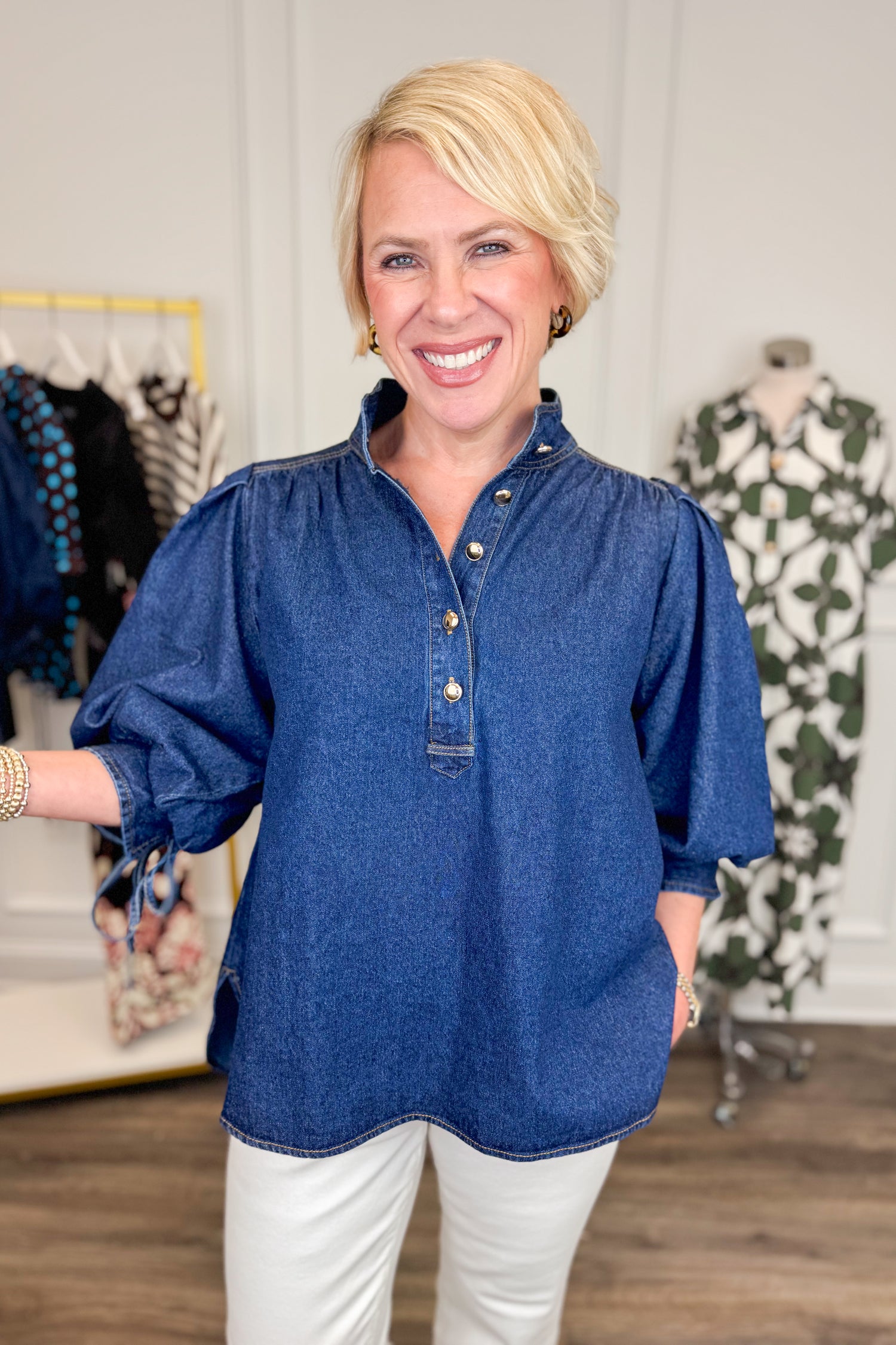 Front view of woman wearing Swagger Boutique’s Dark Wash Puff Sleeve Denim Top by Entro with jeans with clothing hanging in the background.
