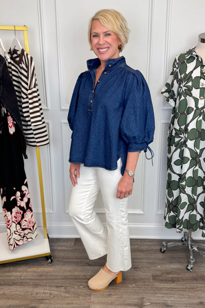 Side view of woman wearing Swagger Boutique’s Dark Wash Puff Sleeve Denim Top by Entro with jeans with clothing hanging in the background.