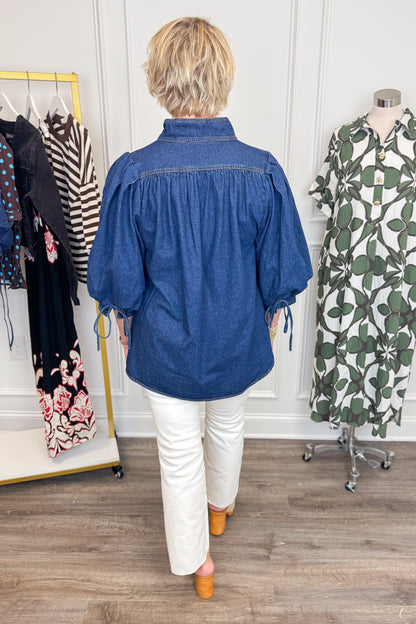 Back view of woman wearing Swagger Boutique’s Dark Wash Puff Sleeve Denim Top by Entro with jeans with clothing hanging in the background.