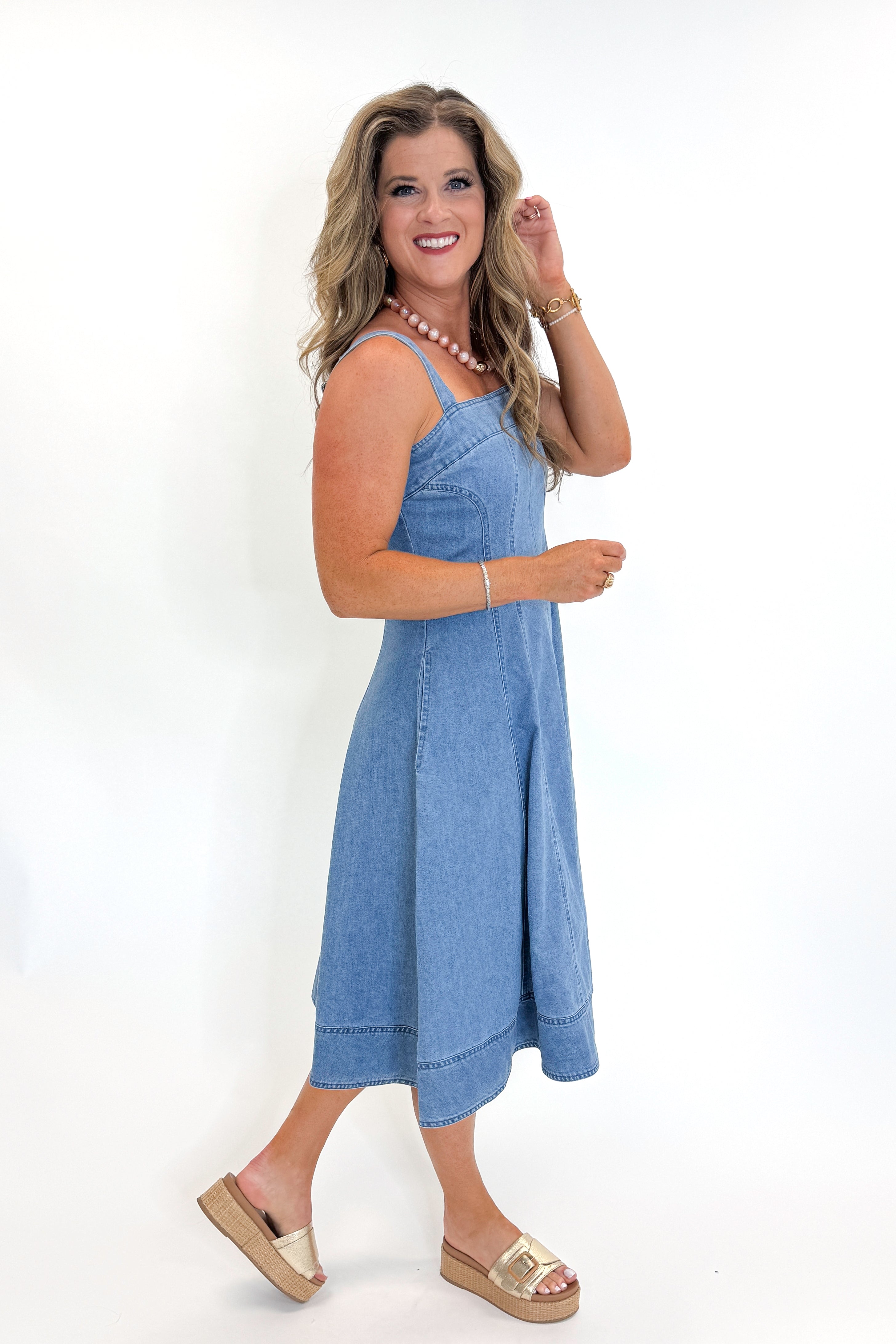 Side view of woman wearing Swagger Boutique’s Denim Fit &amp; Flare Midi Dress with jeans sandals white studio background.