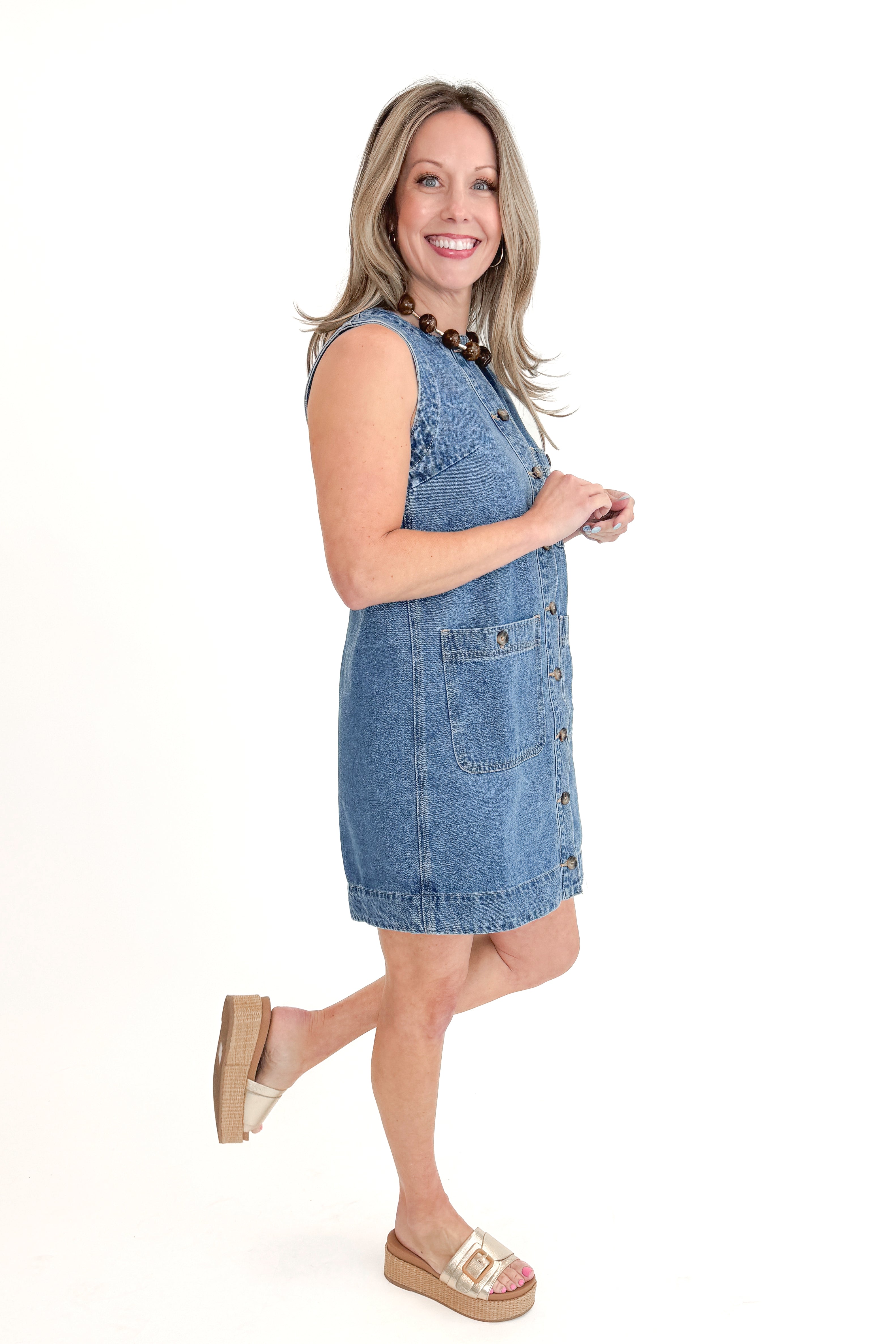 Side view of woman wearing Denim Pocket Detail Dress with sandals against white studio background.