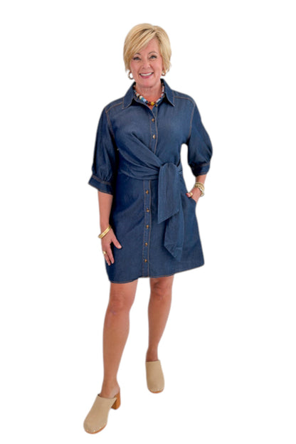 Front view of woman wearing Swagger Boutique’s Denim Tie Detail Button Up Dress by Entro with clogs against white studio background.