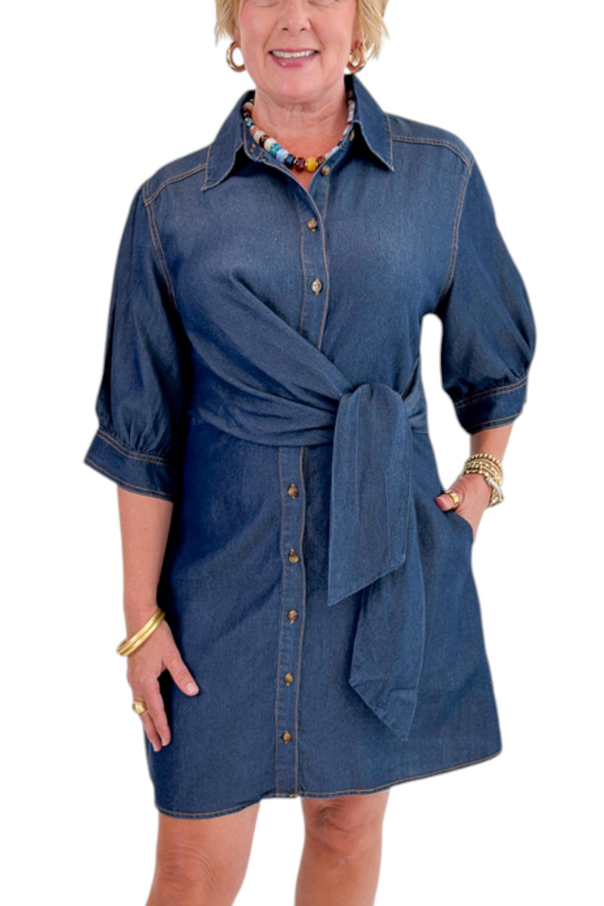 Front detail view of woman wearing Swagger Boutique’s Denim Tie Detail Button Up Dress by Entro with clogs against white studio background.
