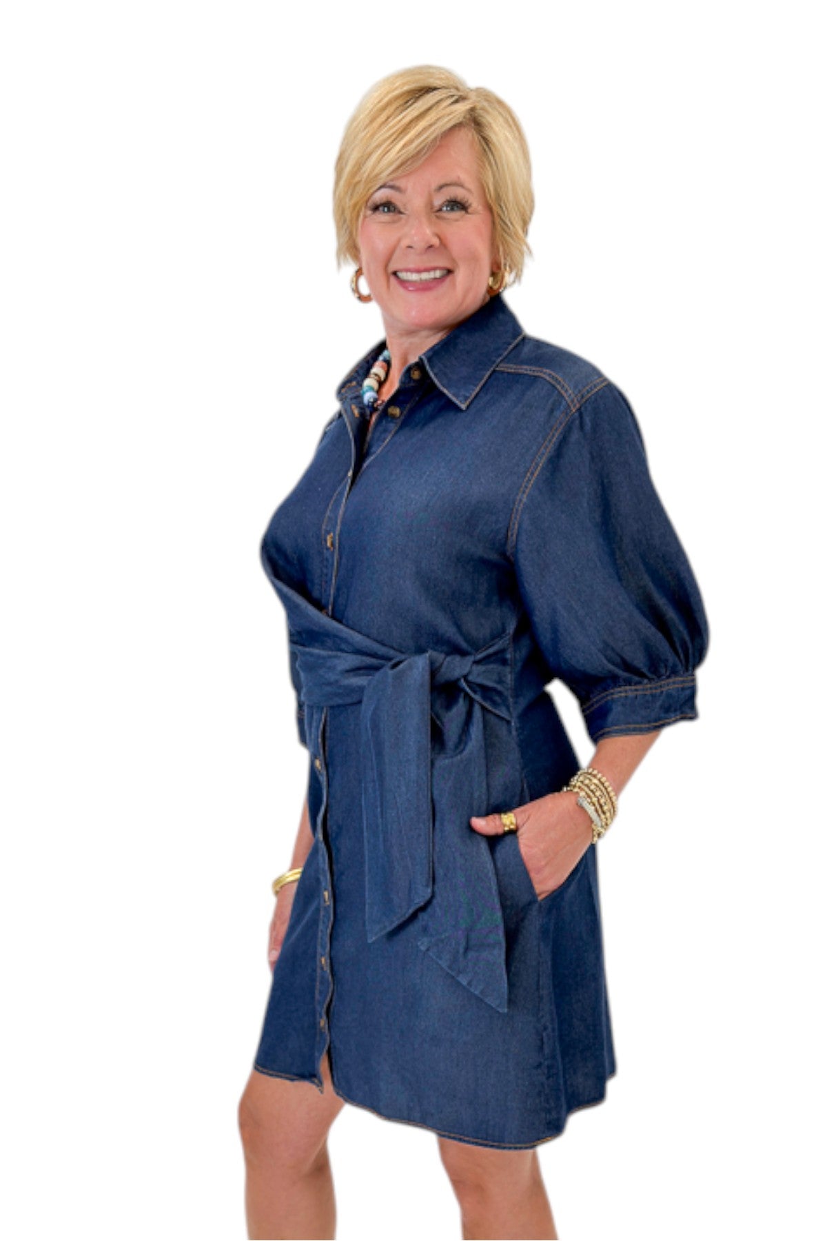 Side view of woman wearing Swagger Boutique’s Denim Tie Detail Button Up Dress by Entro with clogs against white studio background.