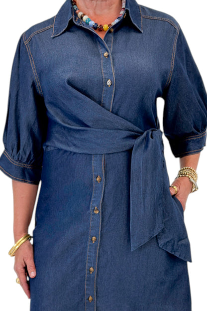 Front detail view of woman wearing Swagger Boutique’s Denim Tie Detail Button Up Dress by Entro with clogs against white studio background.