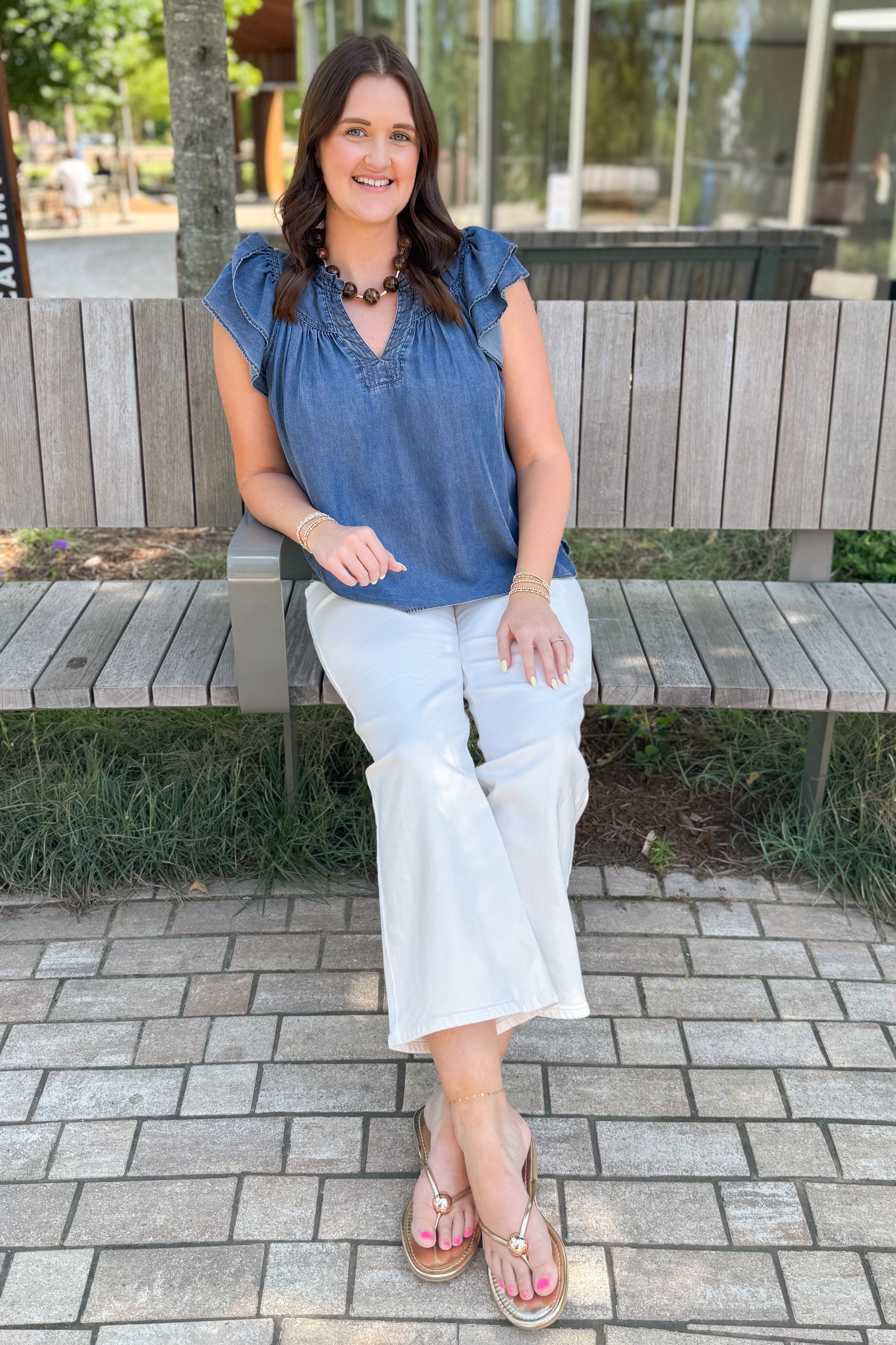 Front view of woman wearing Swagger Boutique’s Denim Ruffle Sleeve Top with jeans sitting on a bench outside.