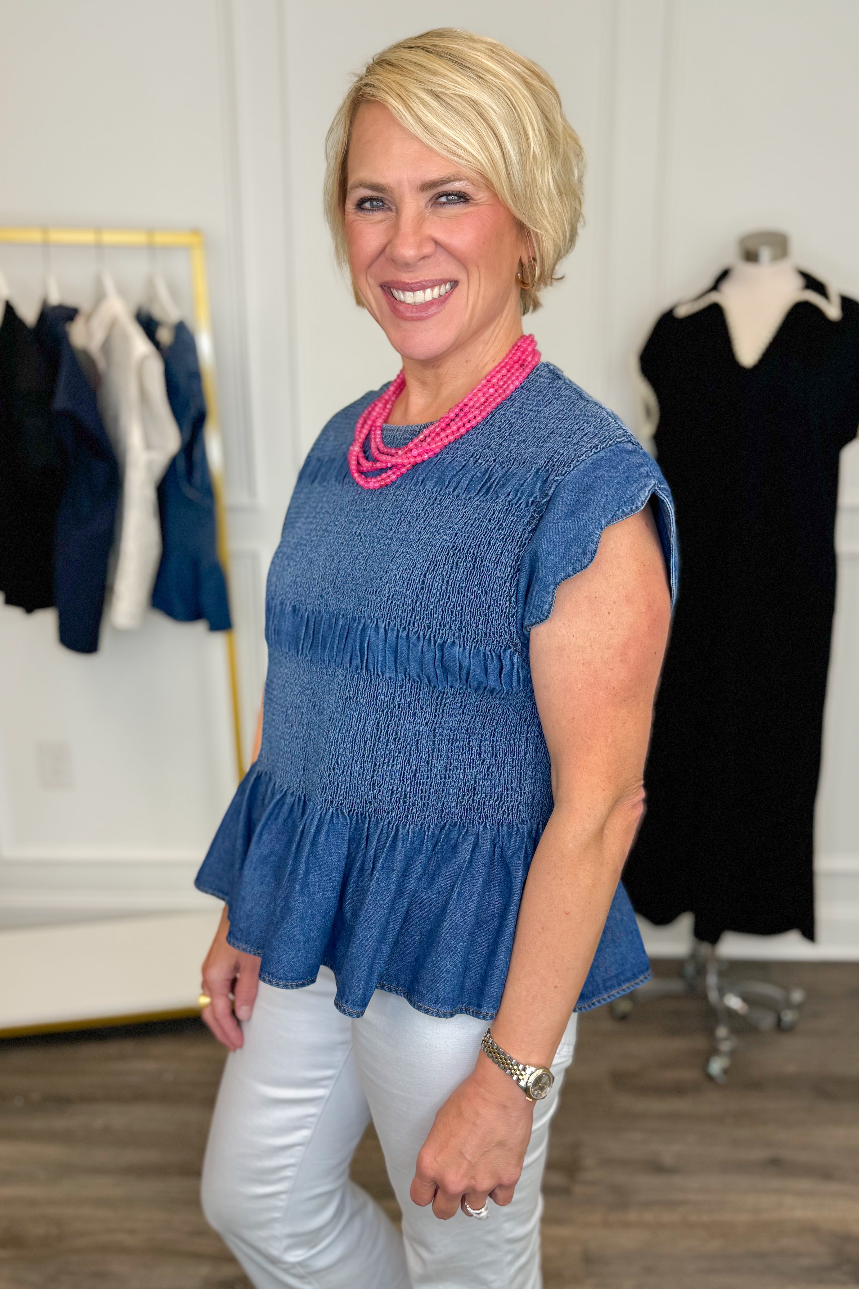 Side view of woman wearing Swagger Boutique’s Denim Smocked &amp; Scalloped Top with jeans with clothing hanging in the background. 