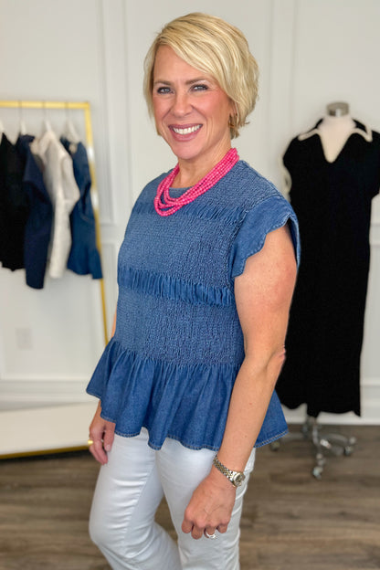 Side view of woman wearing Swagger Boutique’s Denim Smocked &amp; Scalloped Top with jeans with clothing hanging in the background. 