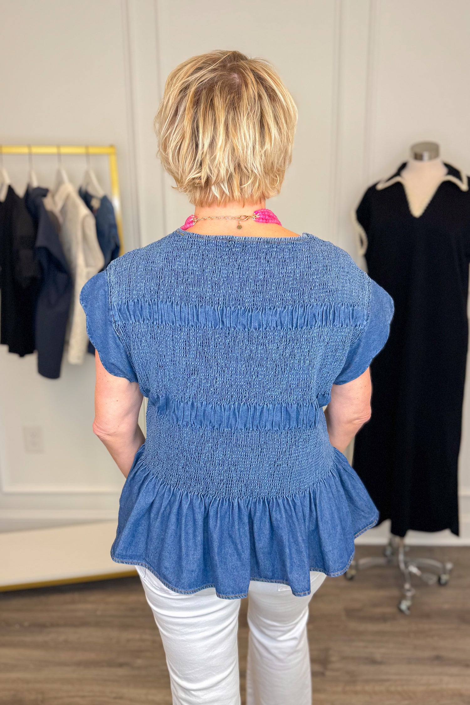 Back view of woman wearing Swagger Boutique’s Denim Smocked &amp; Scalloped Top with jeans with clothing hanging in the background. 