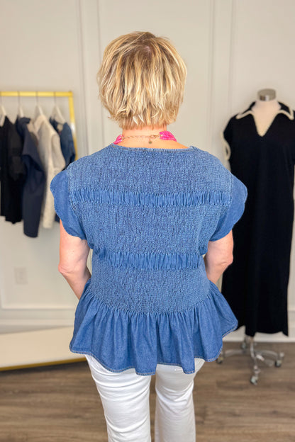 Back view of woman wearing Swagger Boutique’s Denim Smocked &amp; Scalloped Top with jeans with clothing hanging in the background. 