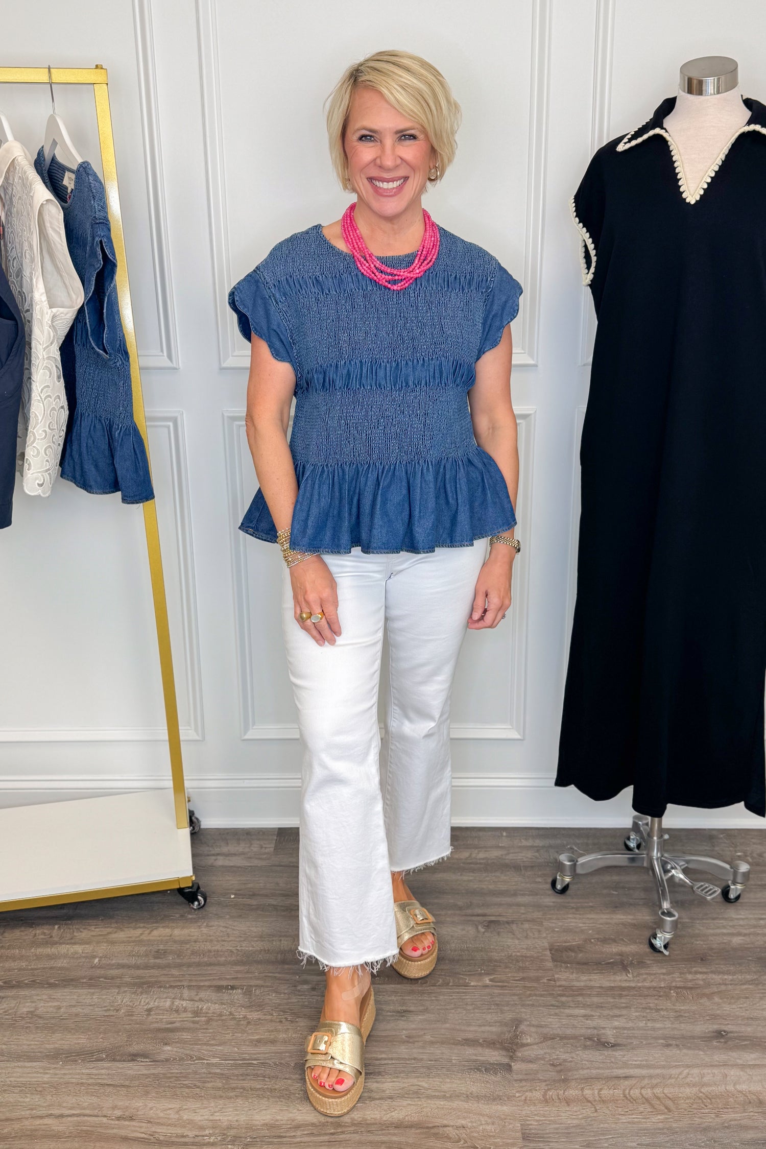 Front view of woman wearing Swagger Boutique’s Denim Smocked &amp; Scalloped Top with jeans with clothing hanging in the background. 