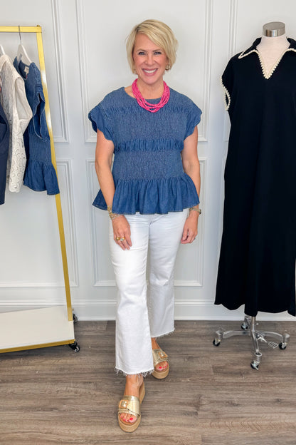 Front view of woman wearing Swagger Boutique’s Denim Smocked &amp; Scalloped Top with jeans with clothing hanging in the background. 