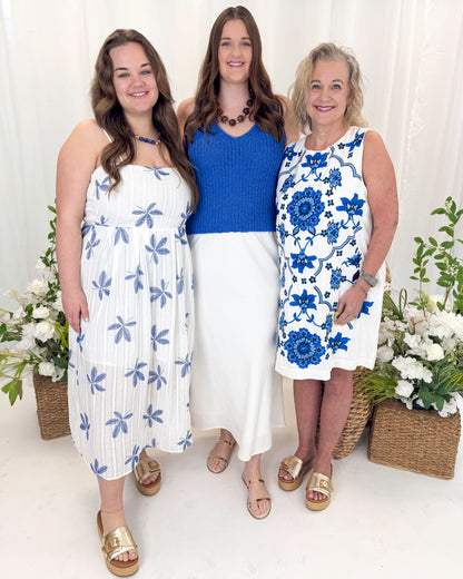 Front view of women wearing Swagger Boutique’s Blue Embroidered Shift Dress by THML, Blue V-Neck Knitted Tank_by Skies Are Blue and White With Blue Embroidery Midi Dress Blue Embroidered Shift with sandals against decorative background.