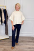 A woman modeling a cream cable-knit turtleneck poncho sweater, paired with dark blue jeans and leopard print shoes, standing in a room with a white paneled wall and a clothing rack.