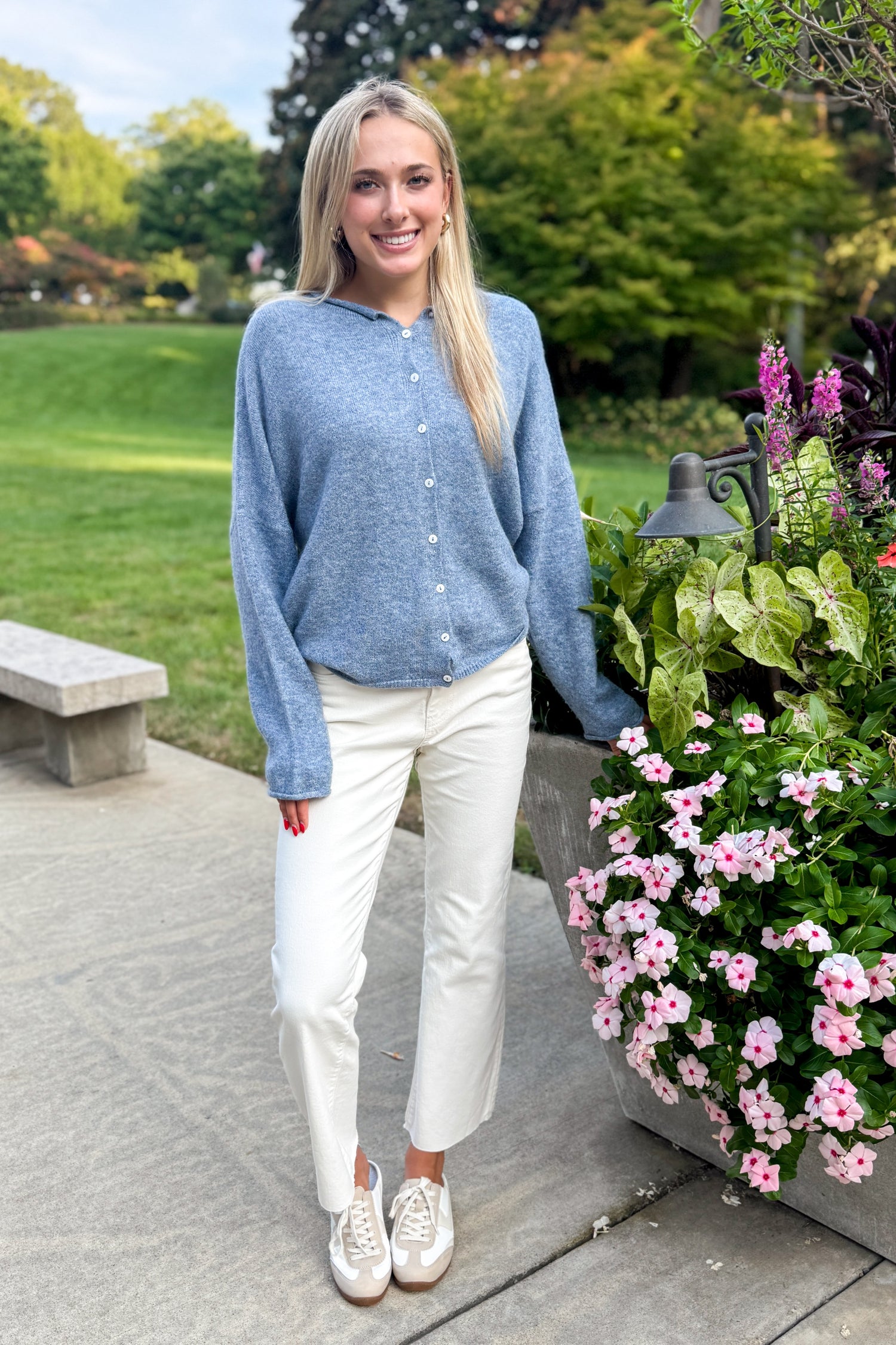 Front view of woman wearing Swagger Boutique’s Dusty Blue Button Up Cardigan with jeans standing outside by flowers. 