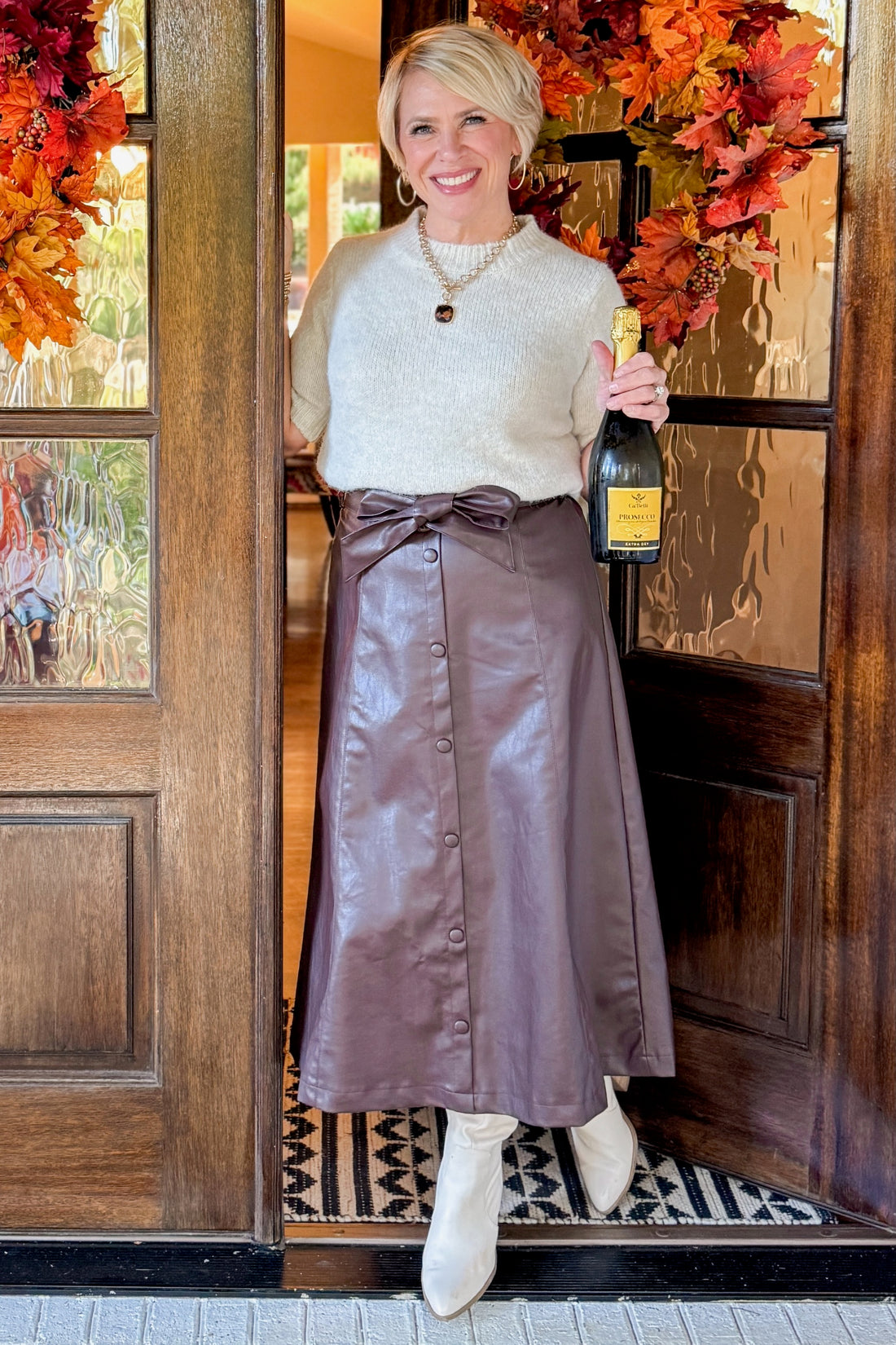 Woman at front door wearing sweater and skirt holding champagne bottle. 