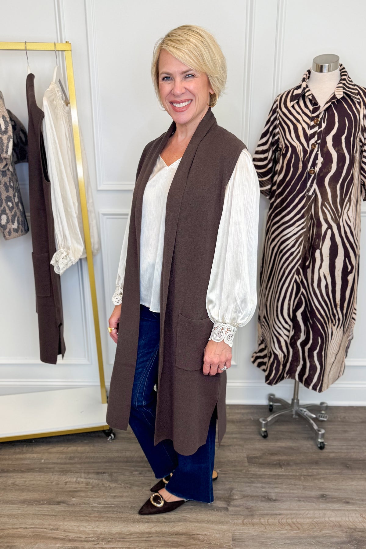 Side view of woman wearing Swagger Boutique’s Chocolate Brown Sleeveless Duster Cardigan with Ivory Satin Cuffed Lace Sleeve Top and Kelsey Dark Wash Denim Jeans with clothing hanging in the background.