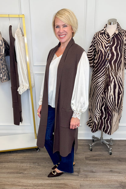 Side view of woman wearing Swagger Boutique’s Chocolate Brown Sleeveless Duster Cardigan with Ivory Satin Cuffed Lace Sleeve Top and Kelsey Dark Wash Denim Jeans with clothing hanging in the background.