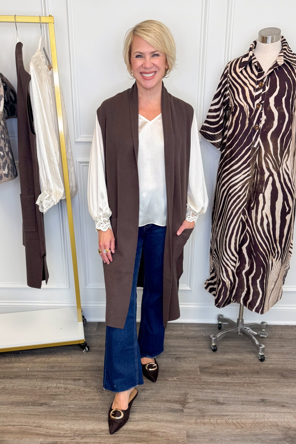Front view of woman wearing Swagger Boutique’s Chocolate Brown Sleeveless Duster Cardigan with Ivory Satin Cuffed Lace Sleeve Top and Kelsey Dark Wash Denim Jeans with clothing hanging in the background.