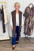 Front view of woman wearing Swagger Boutique’s Chocolate Brown Sleeveless Duster Cardigan with Ivory Satin Cuffed Lace Sleeve Top and Kelsey Dark Wash Denim Jeans with clothing hanging in the background.