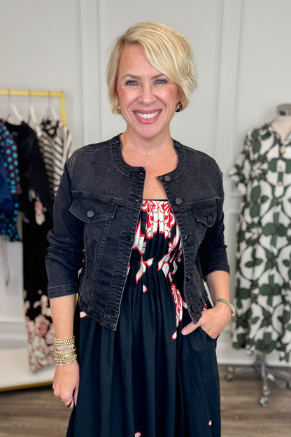 Front view of woman wearing Swagger Boutique’s washed black denim jacked with dress with clothing hanging in the background.