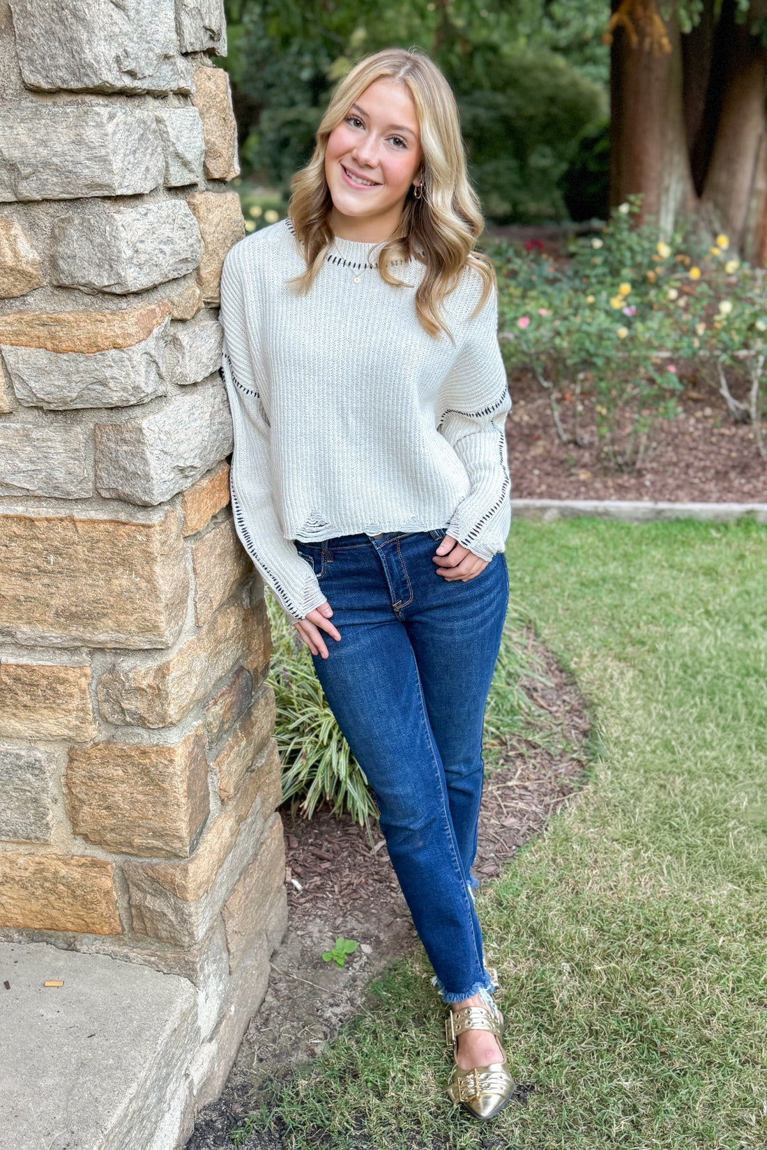 Outside front view of woman wearing stitch detail oatmeal sweater leaning against stone.