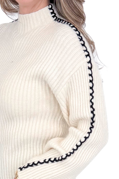 Side detailed view of woman wearing cream sweater with jeans against white studio background.
