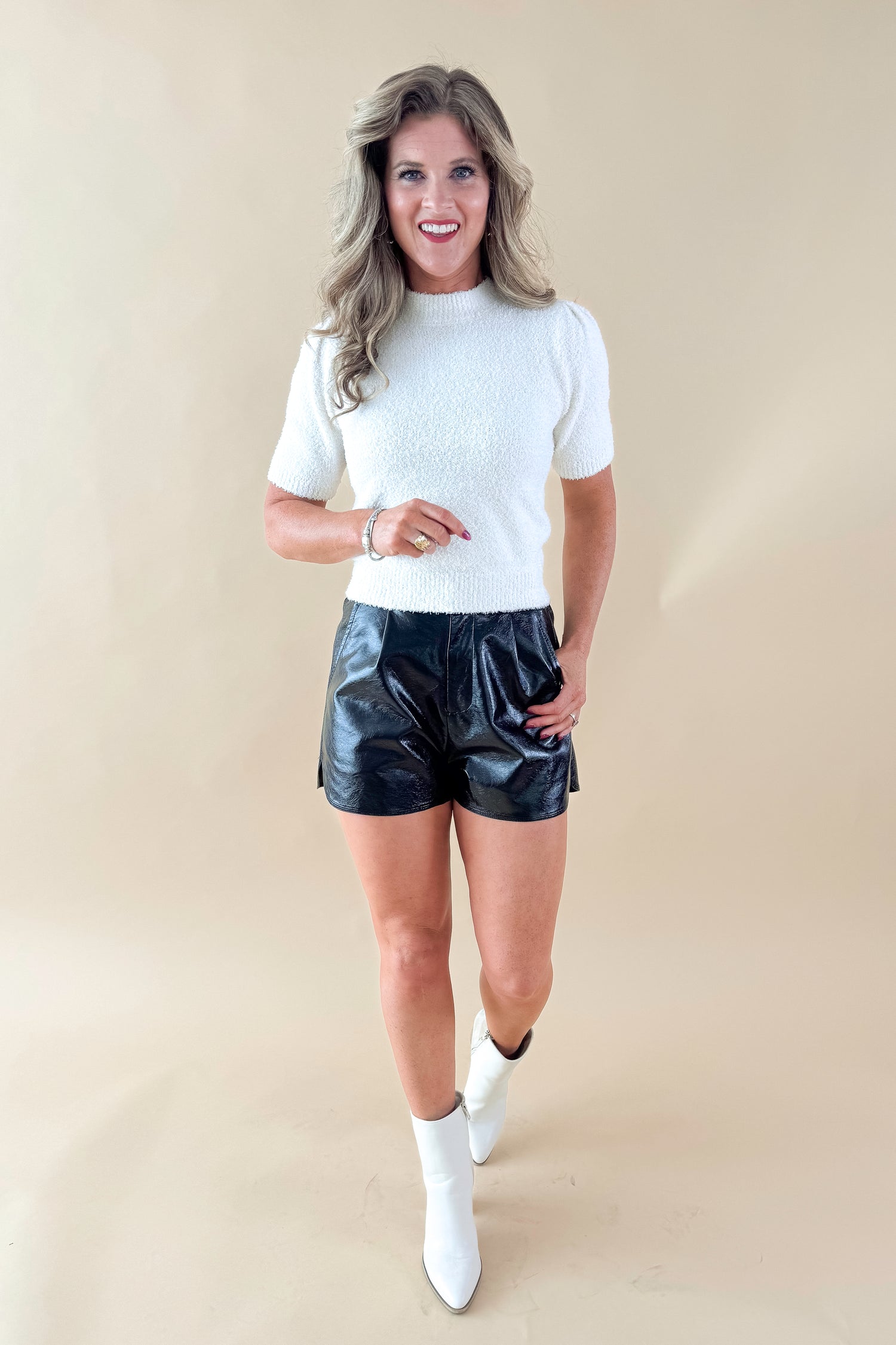 Front view of woman wearing cream sweater with Black Faux Leather Shorts against tan studio background.