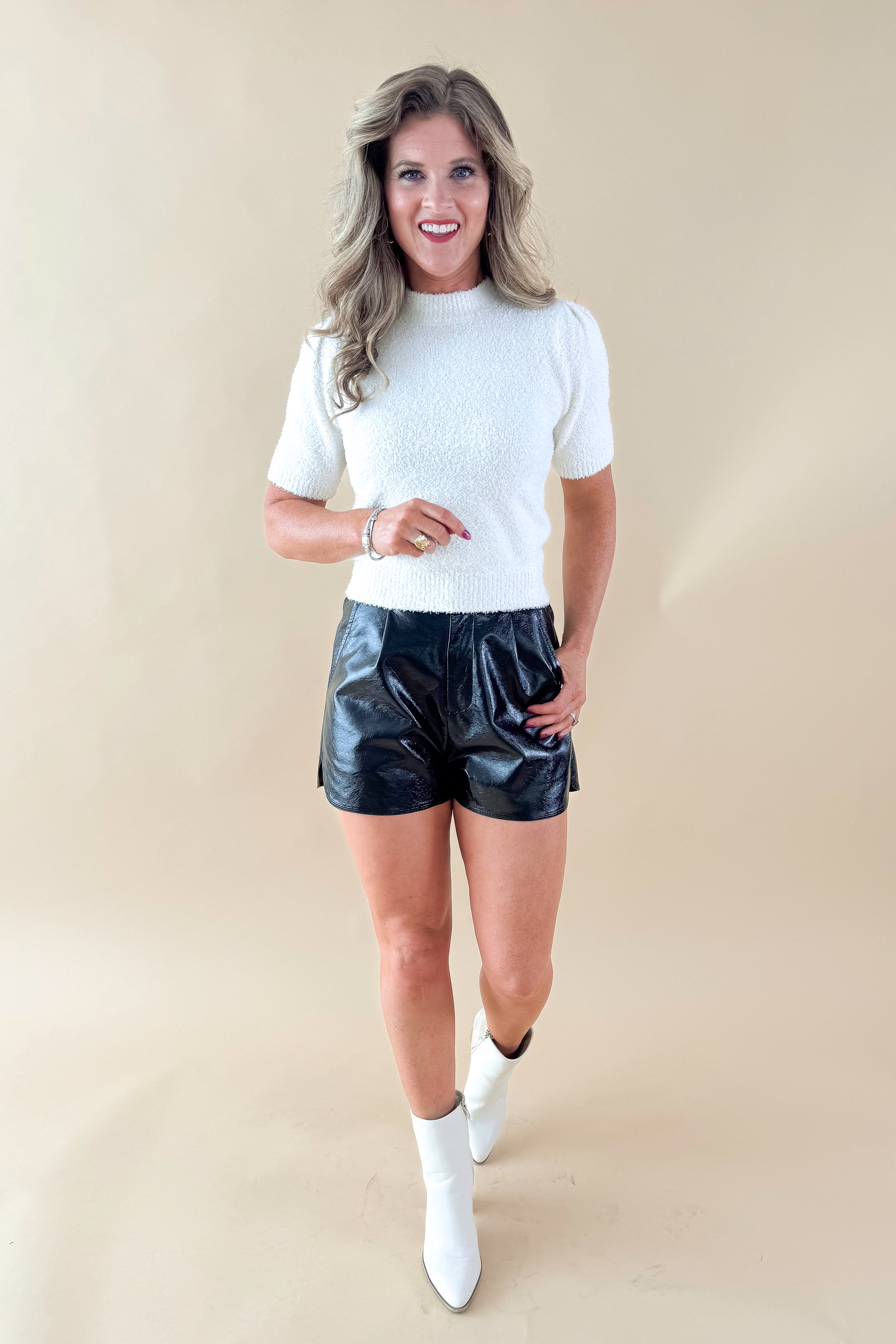 Front view of woman wearing cream sweater with Black Faux Leather Shorts against tan studio background.