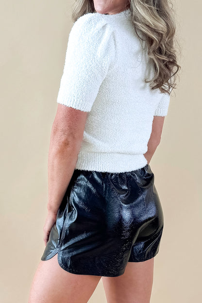 Side view of woman wearing cream sweater with Black Faux Leather Shorts against tan studio background.