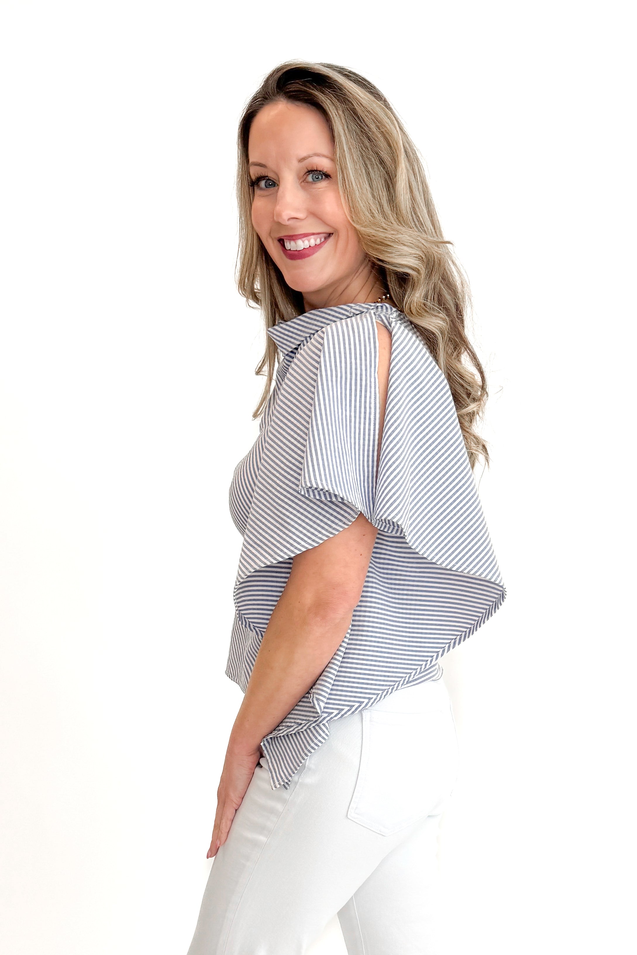 Side view of woman wearing Navy &amp; Ivory Striped Asymmetrical Ruffle Top with jeans and gold bar sandals against white studio background.