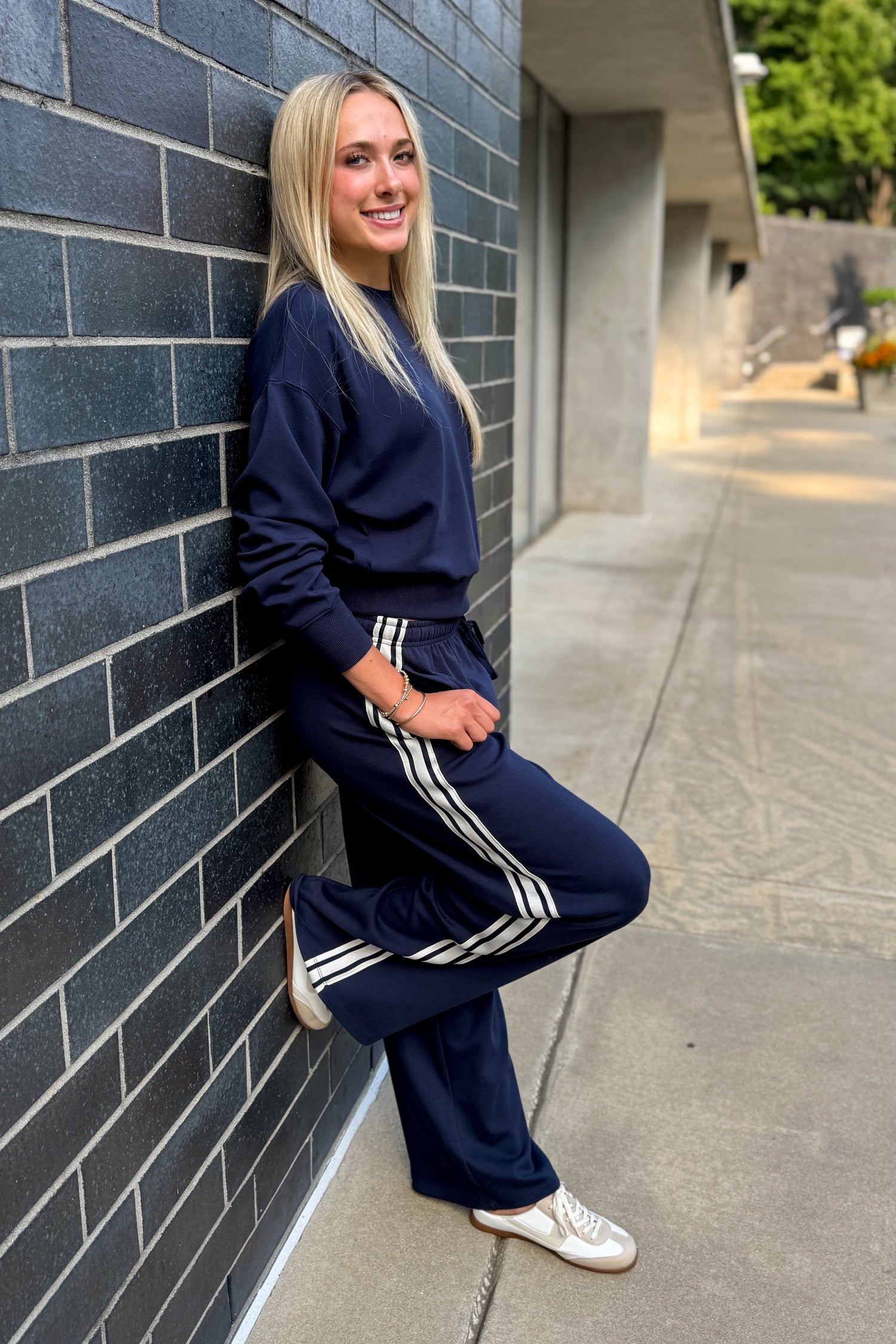 Front view of woman wearing Swagger Boutique’s Eclipse Breakaway Track Pants by Z Supply with Eclipse Comeback Sweatshirt by Z Supply leaning against a brick wall outside.