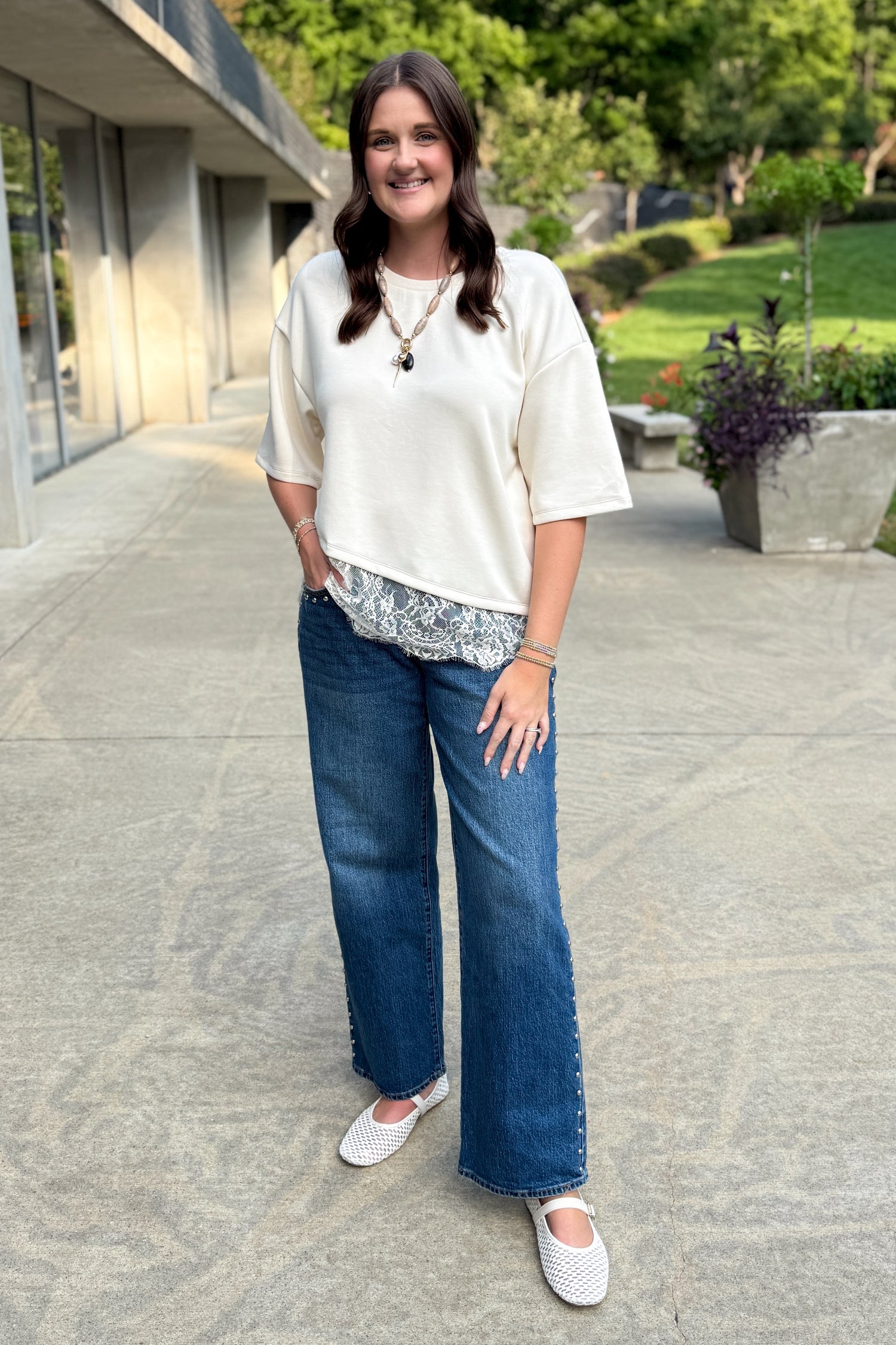 Front view of woman wearing Swagger Boutique’s Ecru Asymmetrical Lace Hem Top by Entro with jeans standing outside.