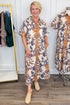 A woman stands in a boutique wearing a short-sleeve button-front midi dress with a cream base and a bold blue and mustard-orange botanical print. A mannequin on the right shows the same dress, and a clothing rack with other garments is visible on the left.