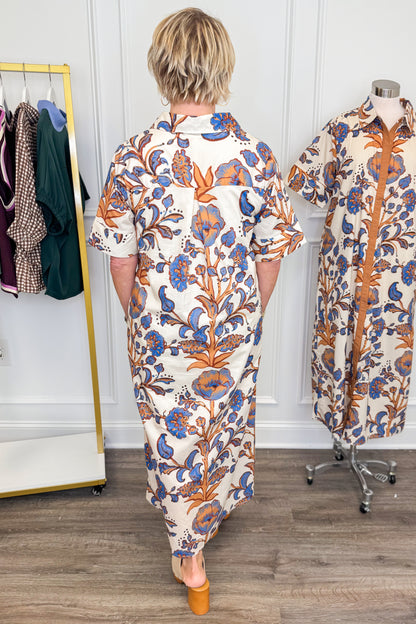 A person viewed from behind wearing a cream shirt dress with a blue and orange floral print, standing in a boutique next to a mannequin displaying the same dress.
